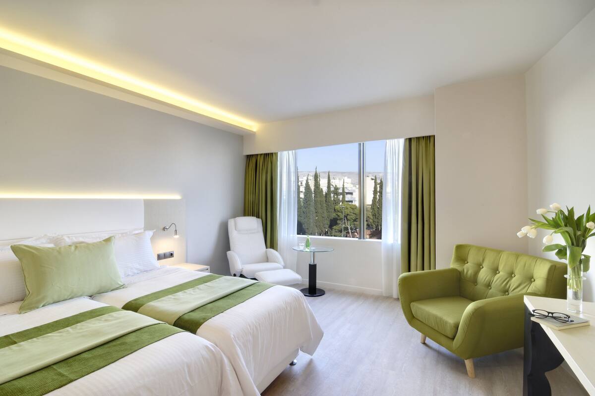 A hotel room with two beds, a green armchair, a small table with a vase, and a view of trees through the window.