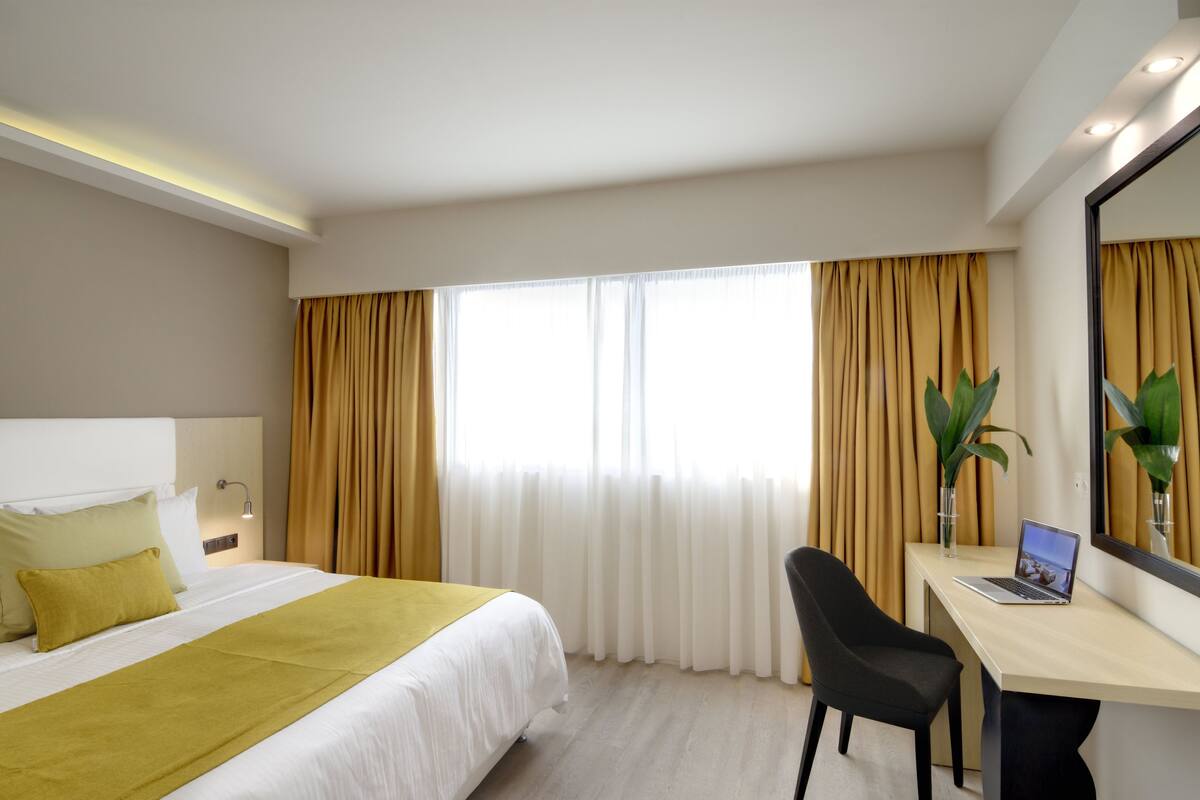 A hotel room with a bed, a desk with a laptop, a chair, a window with curtains, and a mirror.