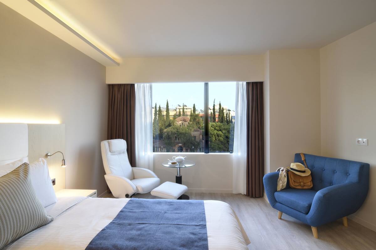 A hotel room with a bed, a white armchair, a blue sofa, a small table, and a view of buildings through large windows.