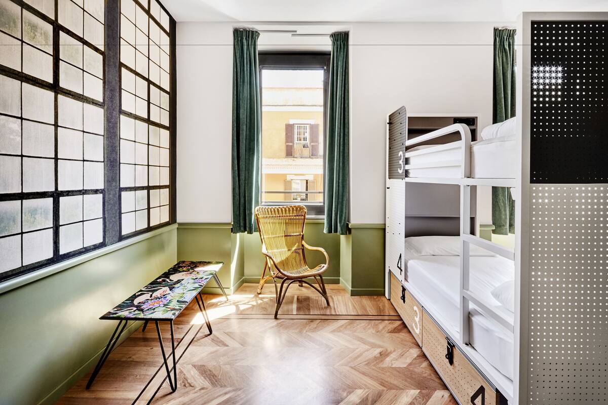 A dormitory room with bunk beds, a wooden floor, a chair, a table, and a window with curtains.