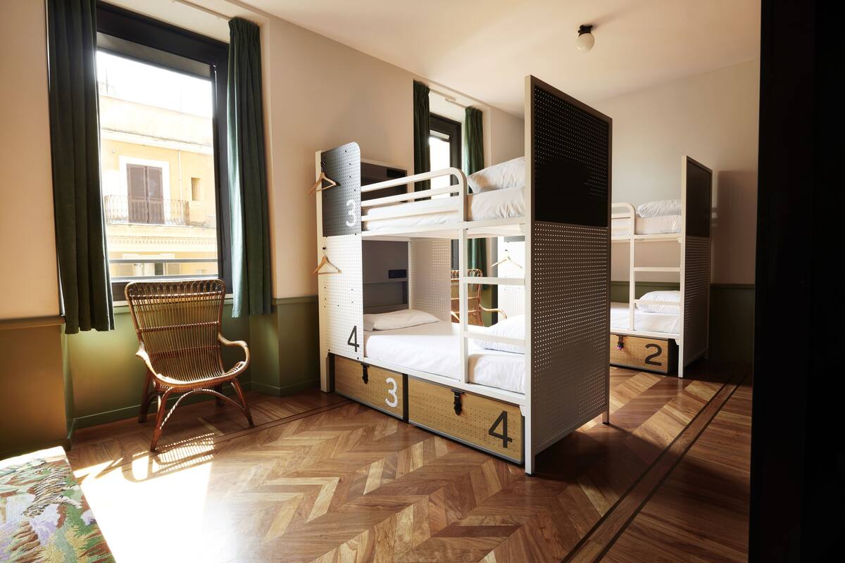 A room with bunk beds, numbered drawers, a chair, and a window with curtains.