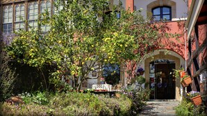 Une cour avec un jardin, une table entourée de chaises et un bâtiment doté de fenêtres en arc de cercle et d’une porte.