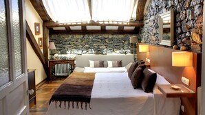 Une chambre avec un mur en pierre, un plafond en bois, un lit recouvert d’une courtepointe blanche et marron, une table de chevet avec une lampe, et un miroir encadré accroché au mur.