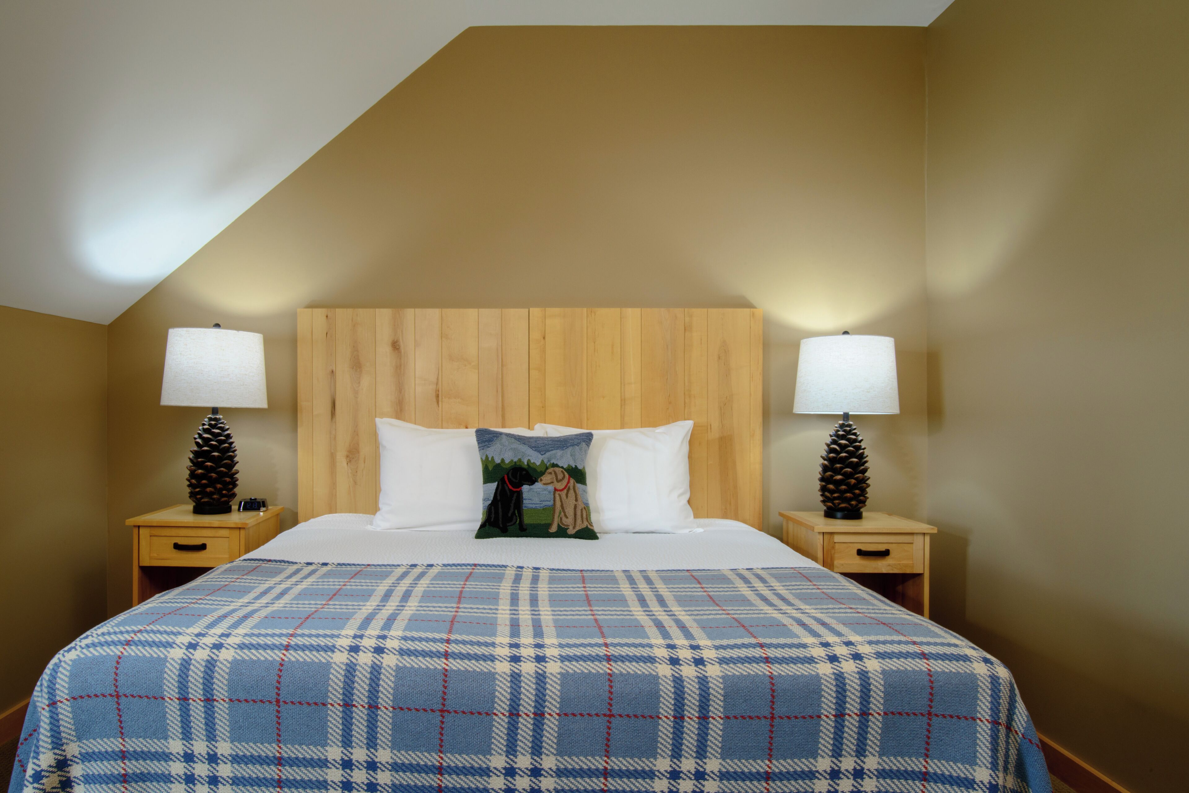 A hotel room with a wooden headboard, two bedside tables with lamps, a plaid bedspread, and a pillow with a dog illustration.