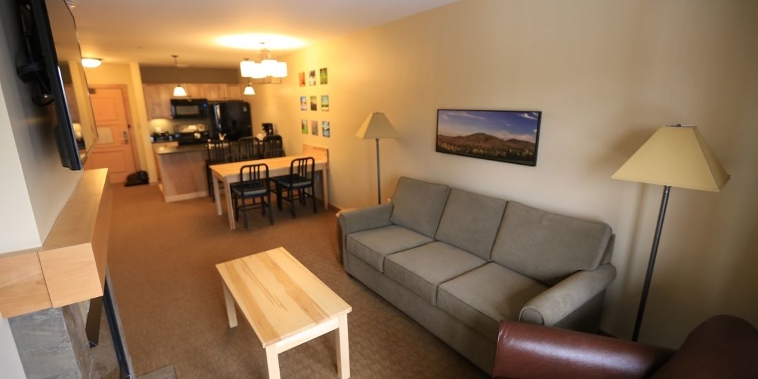 Een woonkamer met een grijze bank, een houten salontafel en een fauteuil van bruin leer. Er is een eethoek met een tafel en stoelen, en een keuken op de achtergrond.