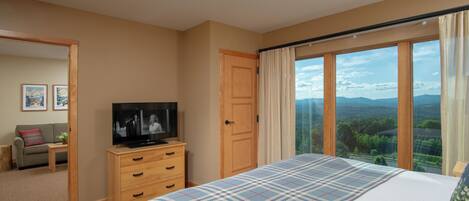 Un dormitorio con una cama grande, un televisor sobre un tocador de madera, una puerta que da a otra habitación y vistas al exterior a través de una ventana.