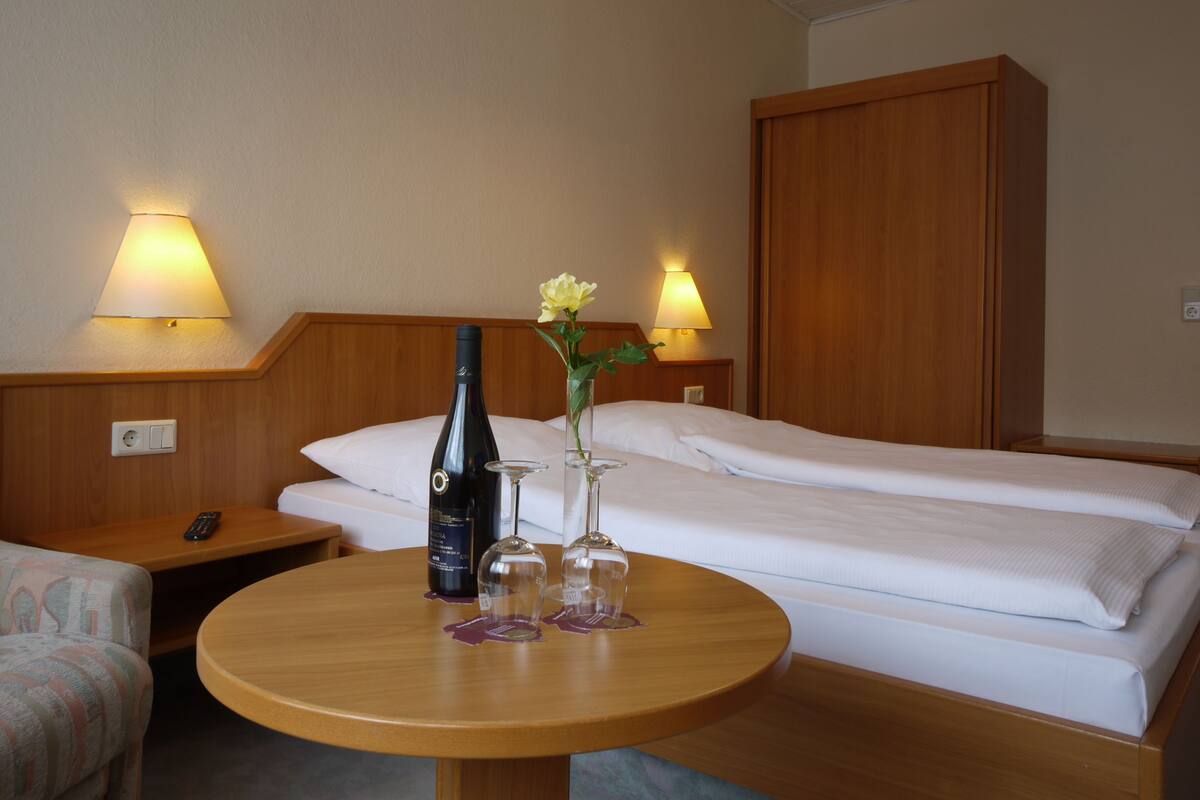 Una habitación de hotel con una cama, una mesita de noche con una botella de vino y dos copas, un control remoto y dos lámparas en la pared.