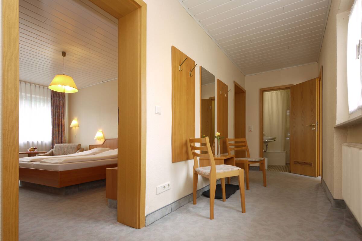 Una habitación de hotel con cama, dos sillas, una mesa, un espejo y un baño visible a través de una puerta abierta.