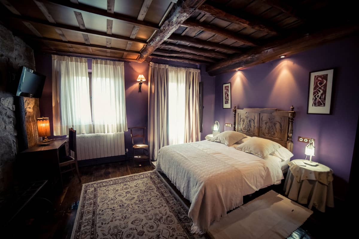 A bedroom with a large bed, a wooden headboard, a nightstand with a lamp, a chair, and a radiator.