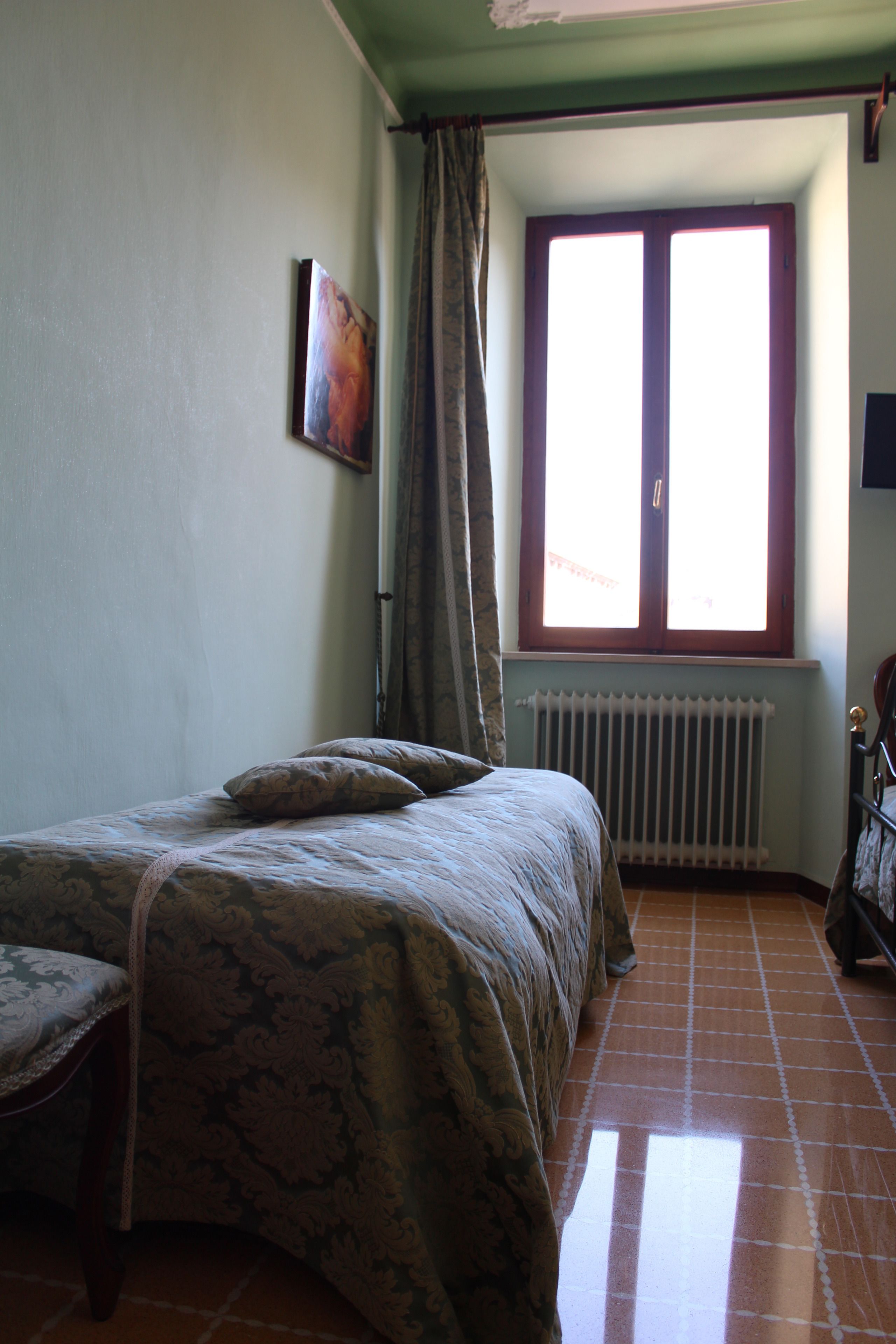 A bedroom with a bed, a radiator, a window with curtains, and a painting on the wall.