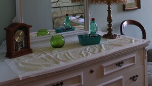 A room with a large ornate mirror, a bedside lamp, a dresser with drawers, a clock, and a framed picture on the wall.