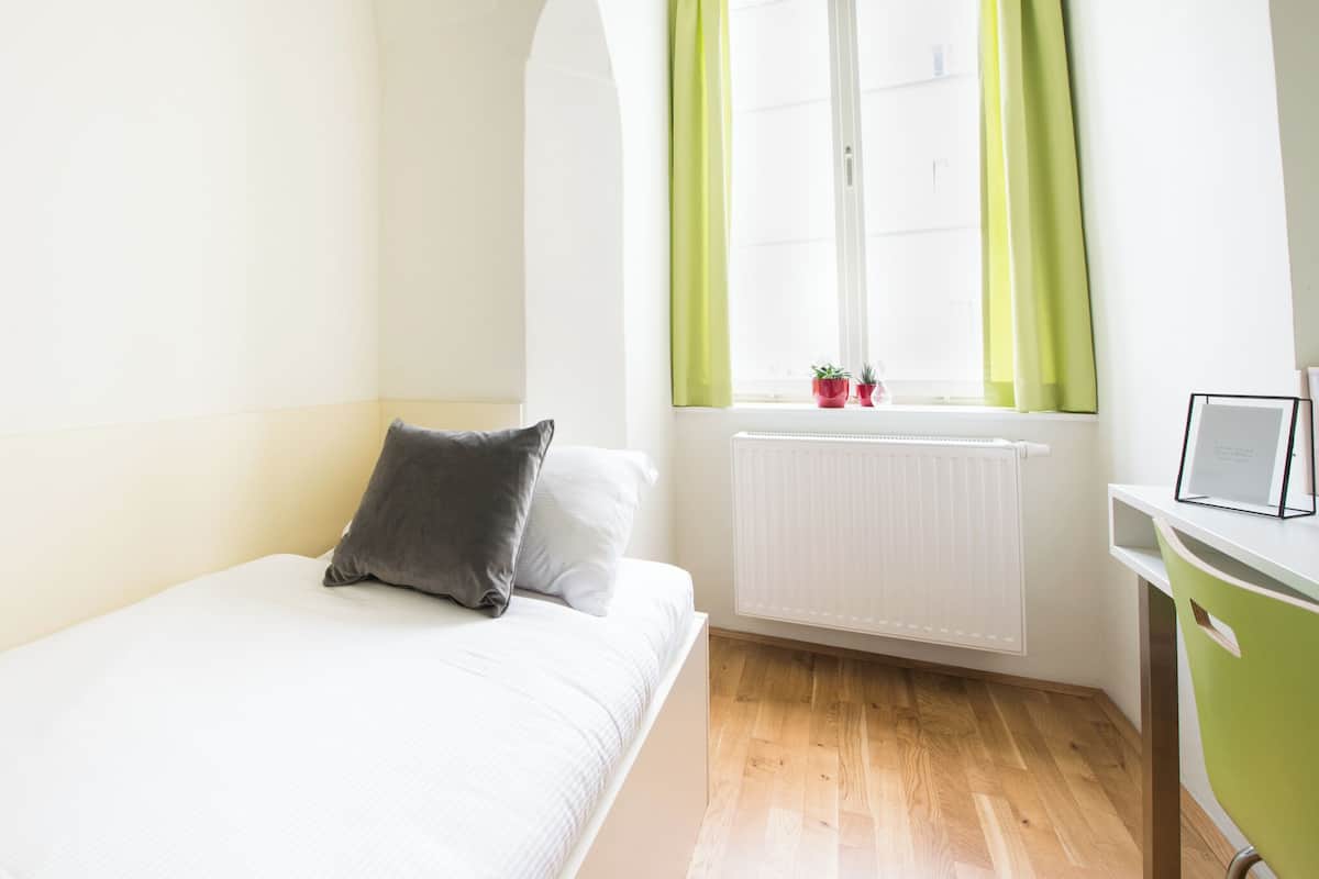 Een kamer met een bed, een raam met gordijnen, een radiator en een bureau met stoel.
