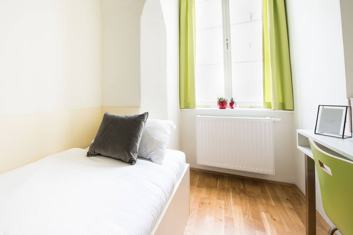 Una camera da letto con un letto, una finestra con tende, un termosifone e una scrivania con una sedia.