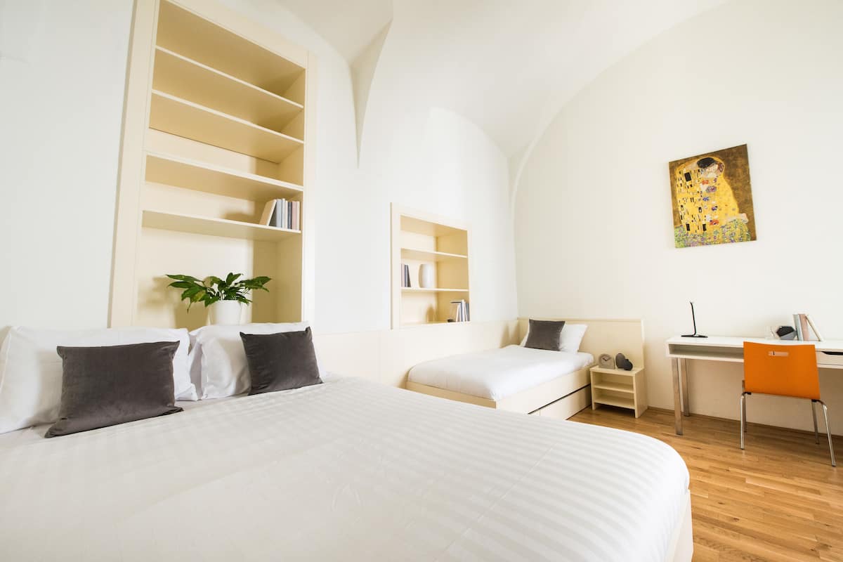 Una camera d'albergo con un letto grande, una scrivania e una sedia. Si notano librerie e un quadro appesi al muro.