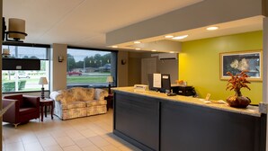 A reception area with a counter, a sofa, a red armchair, and a window with a view of a parking lot and greenery. - Regency Inn (Monroeville)