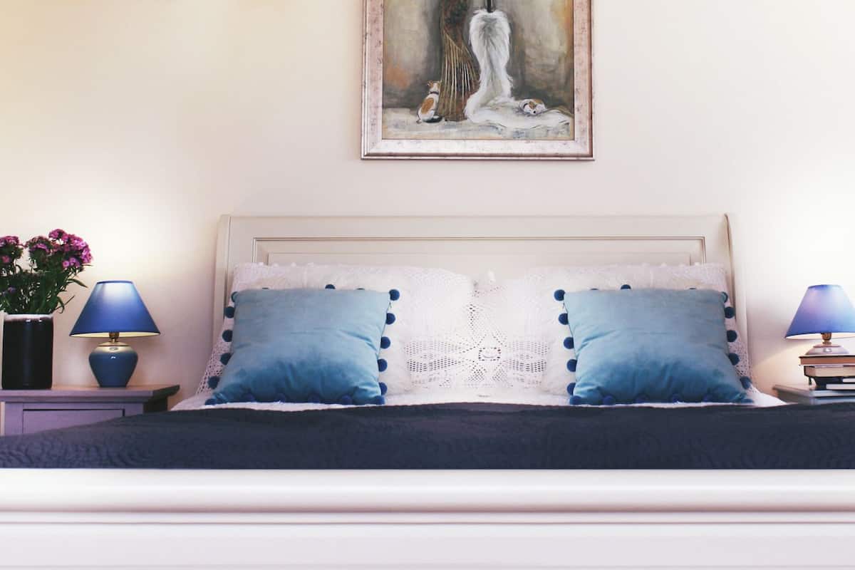 Ein ordentlich bezogenes Bett mit zwei Kissen, ein Nachttisch mit Lampe, eine Vase mit Blumen und ein Bild an der Wand.