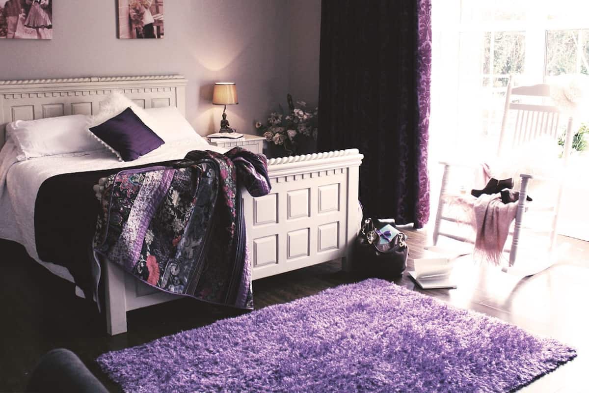 Ein Schlafzimmer mit einem weißen Bett, violetter Bettwäsche, einem Nachttisch mit Lampe und einem Fenster mit Vorhängen.