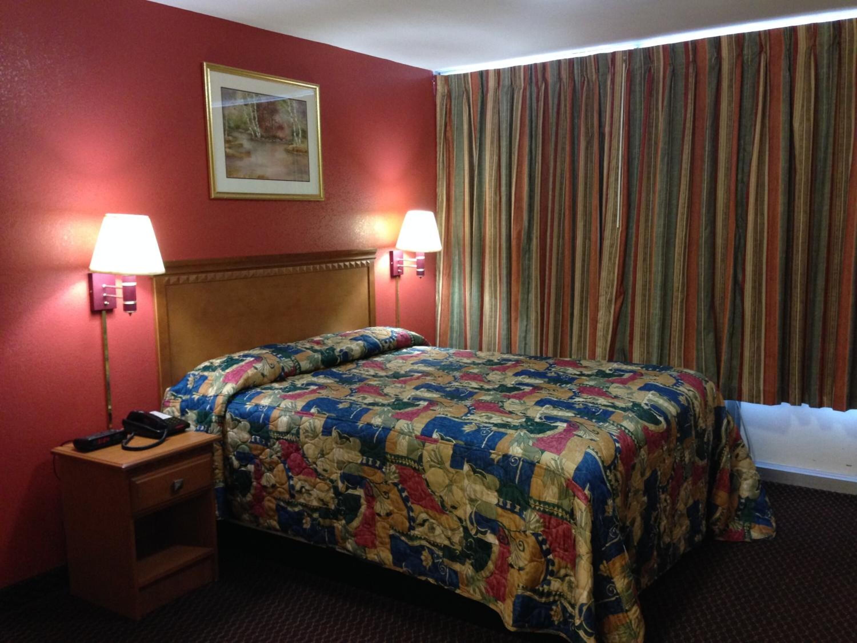 Una habitación de hotel con cabecera de madera, cubrecama estampado con flores, dos lámparas de noche, una mesita de noche y una ventana con cortinas a rayas.