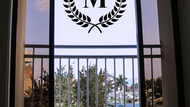 A glass door with a logo featuring a 'M' inside a laurel wreath and two stars above, set against a view of a cityscape with buildings and trees. - Musky Hs & Suites (Chiclayo)