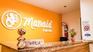 Un comptoir d’accueil avec une enseigne indiquant « SORRIA » et une figurine décorative. Un extincteur et un panneau « interdiction de fumer » sont visibles sur le mur.