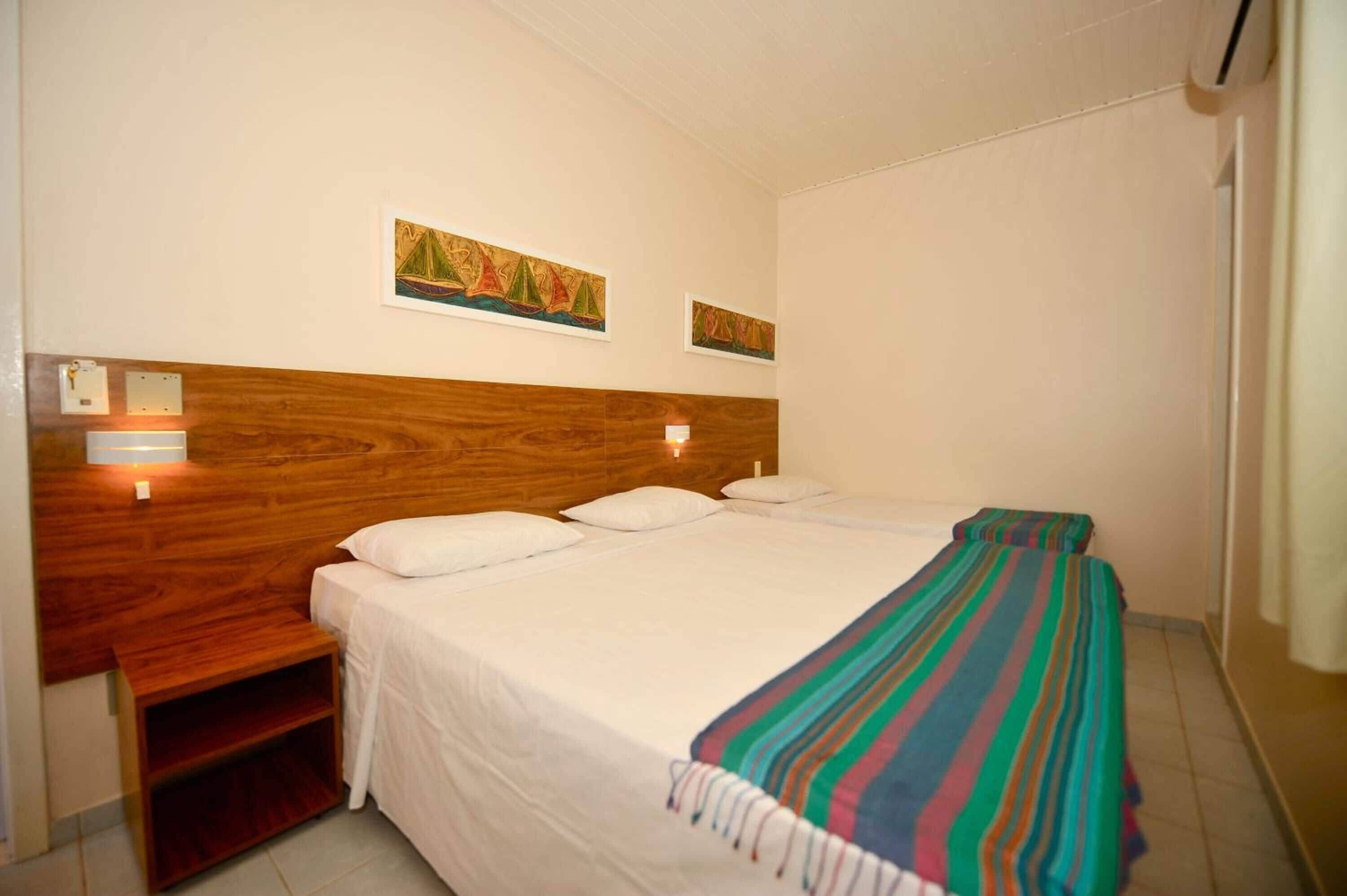 Une chambre d’hôtel avec une tête de lit en bois, deux lits, une couverture rayée aux couleurs vives et des lampes fixées au mur.