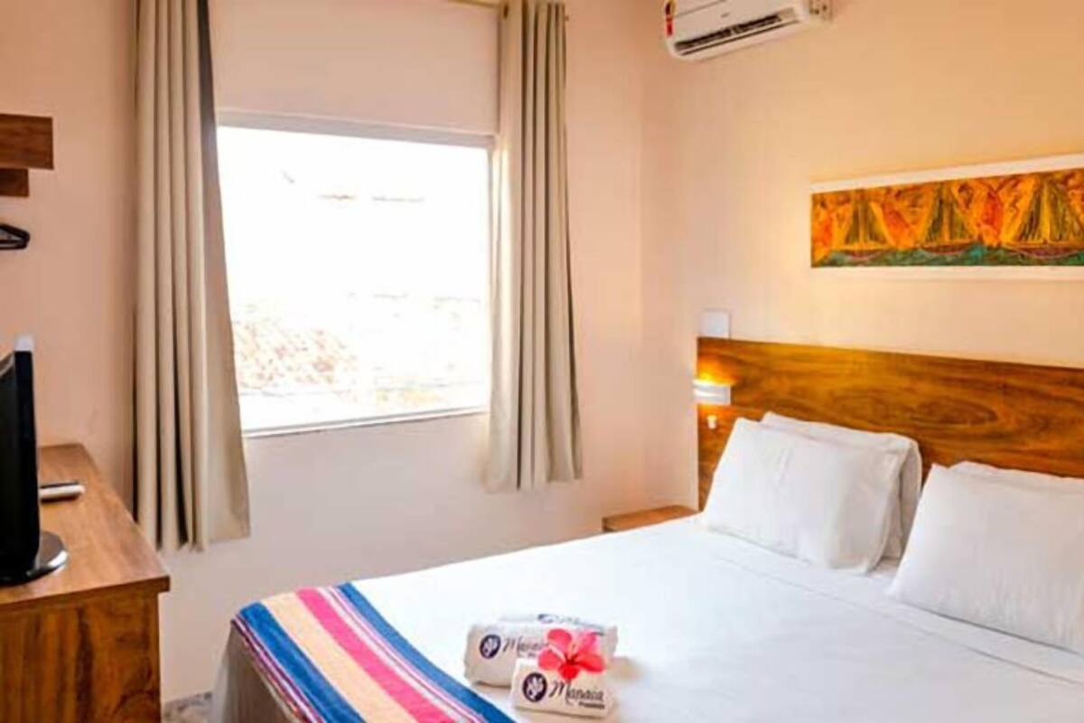 Une chambre d’hôtel avec un lit, un téléviseur posé sur une table en bois, une fenêtre avec des rideaux et un tableau accroché au mur.