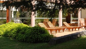 A patio area with wooden lounge chairs and a table, surrounded by greenery and a stone wall.