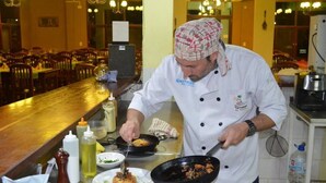 A chef in a white uniform with a patterned headscarf preparing food in a kitchen with a long counter and multiple chairs in the background. - Terrazas de Colon (Colon)
