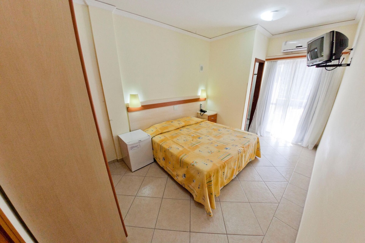 A hotel room with a bed, bedside tables, a lamp, a TV, and an air conditioning unit.