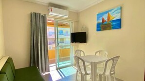 A room with a green sofa, a dining table with white chairs, a flat-screen TV mounted on the wall, and a painting of sailboats. - Bombinhas Praia Apart Hotel - Unidade Bem Te Vi (Bombinhas)