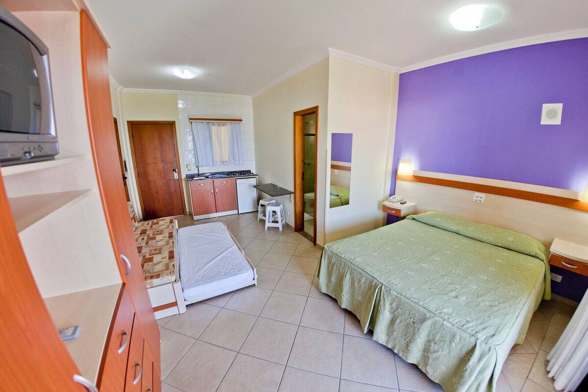 A compact hotel room with a bed, a small kitchenette, a bathroom, and a TV.