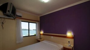 A hotel room with a bed, a nightstand, a lamp, a TV, and a window with a view of the sea. - Bombinhas Praia Apart Hotel - Unidade Bem Te Vi (Bombinhas)