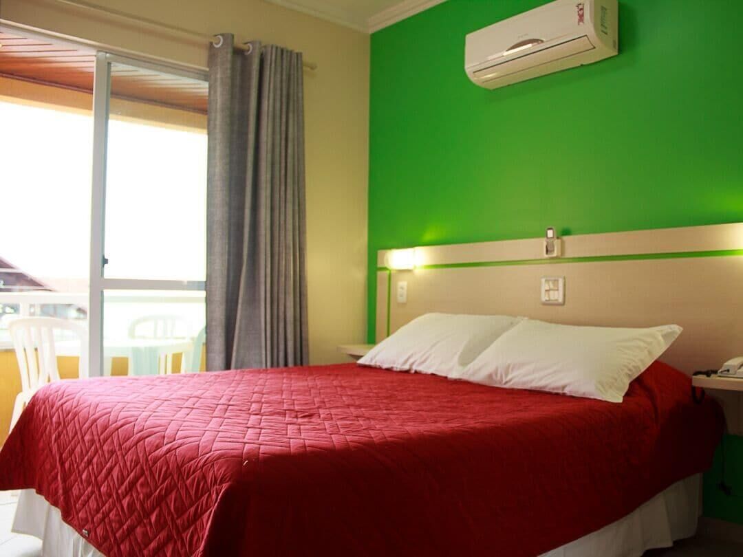 Quarto de hotel com colcha vermelha, parede verde e porta de correr para a varanda.