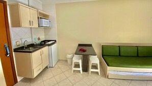 A compact living space with a kitchenette, a dining table with two stools, and a green sofa. - Bombinhas Praia Apart Hotel - Unidade Bem Te Vi (Bombinhas)