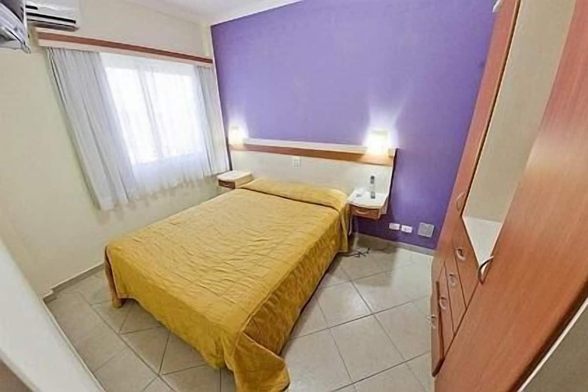 A hotel room with a bed, a nightstand, a window with curtains, and a purple accent wall.