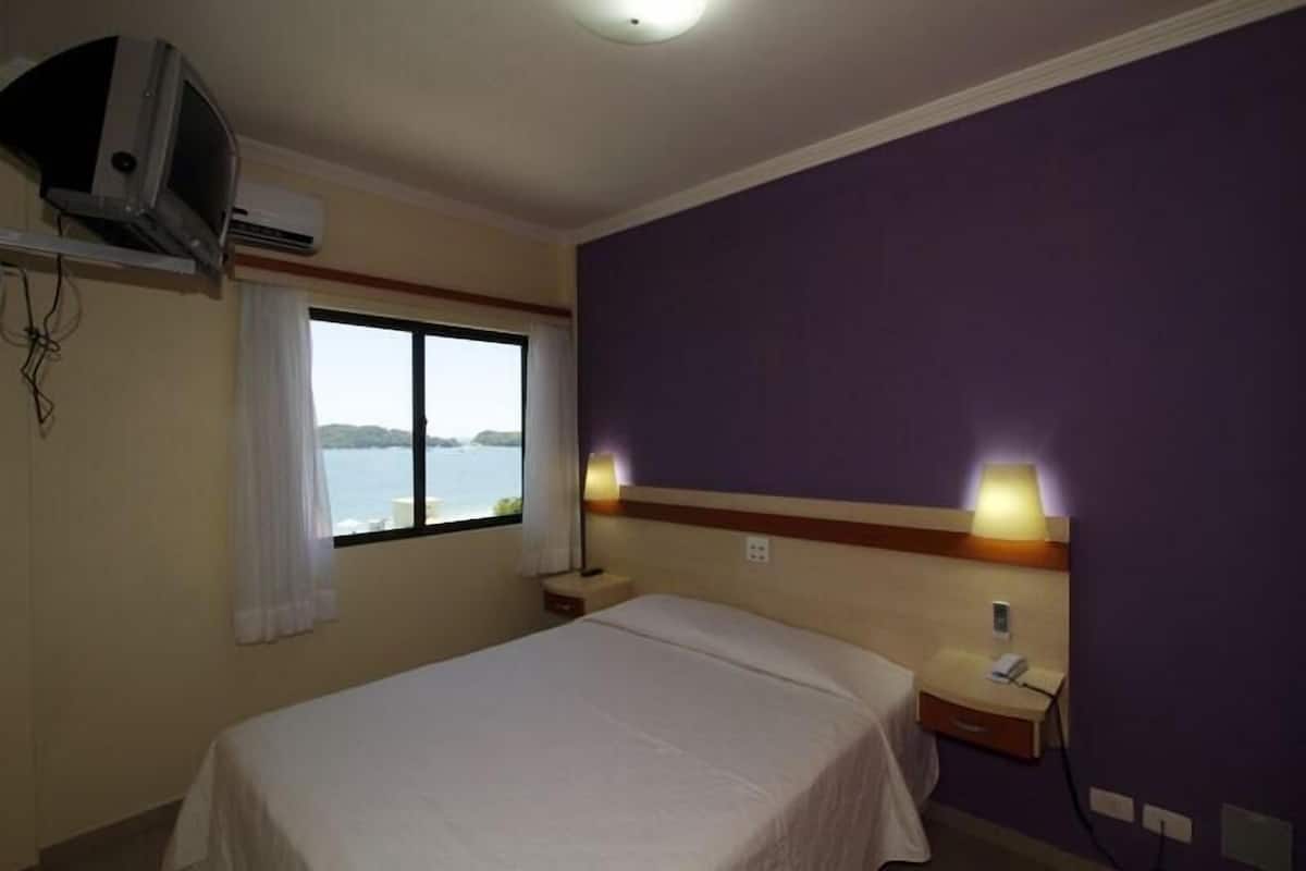 A hotel room with a bed, a nightstand, a lamp, an air conditioner, and a window with a view of the water.