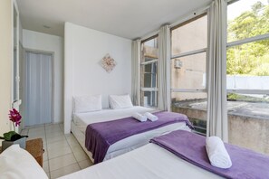 Two beds with crisp linens and comfortable bedspreads, a potted plant, and a large window offering a view of the outdoors.