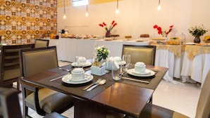 Un comedor con una mesa dispuesta para dos personas, con platos, cuencos y cubiertos blancos. Hay sillas alrededor de la mesa y una mesa de buffet con diversos alimentos al fondo.