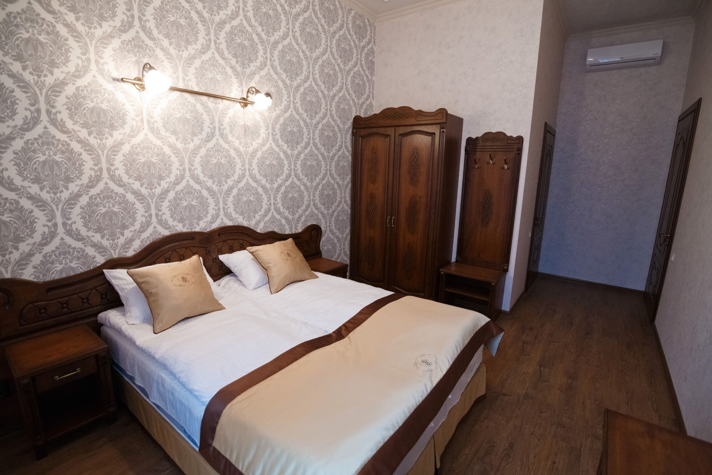 A neatly made bed with a wooden headboard, a nightstand, and a wardrobe in a room with patterned wallpaper and wooden flooring.