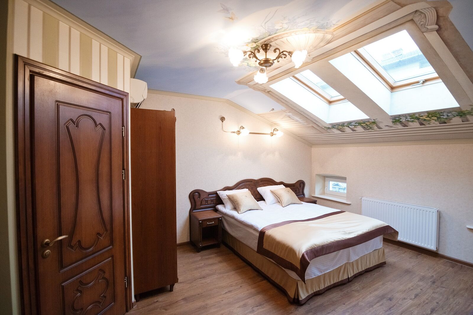 A bedroom with a wooden wardrobe, a bed with a headboard, a nightstand, a wall-mounted light fixture, and skylights.
