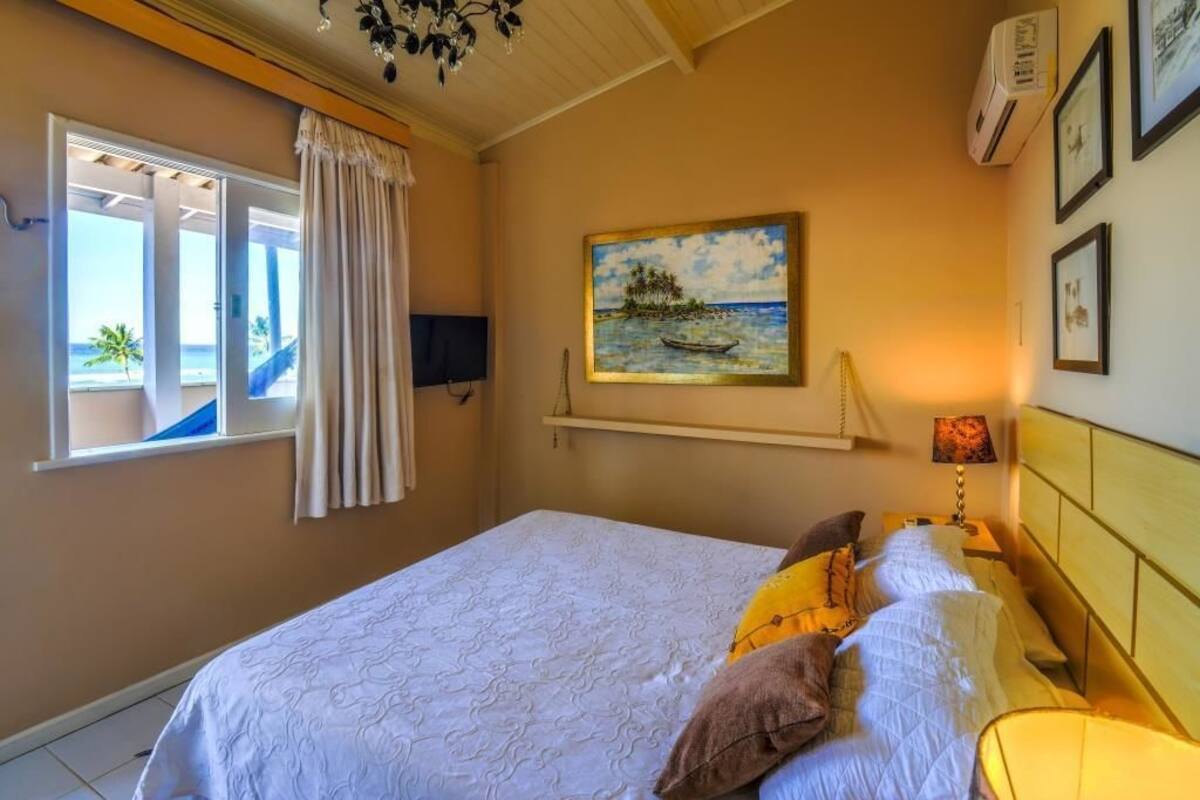 A bedroom with a bed, a painting, a window with curtains, and a wall-mounted air conditioner.
