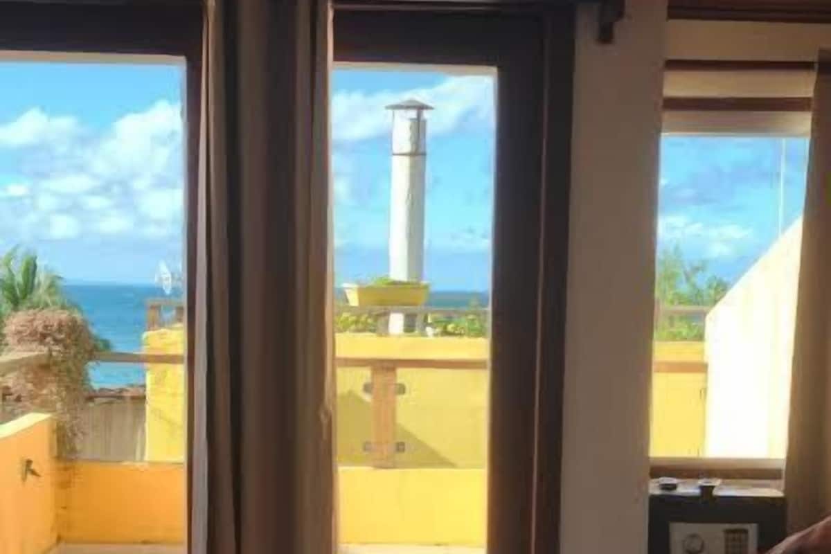 Huoneessa on sänky, ikkunassa verhot ja ikkunasta avautuu näkymä merelle ja rakennuksiin.