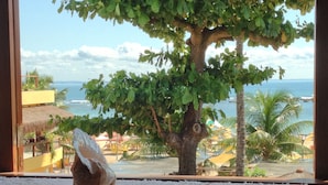 A window view of a beach with a tree, a thatched-roof structure, and a white furry rug on the windowsill. - Pousada Funny (Morro de Sao Paulo)
