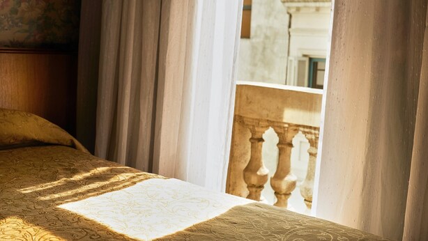 A bed with a patterned bedspread, curtains, and a view of a building through a window with a balustrade. - Gran Hotel Ailen (Buenos Aires)