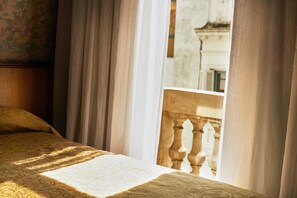 A bed with a patterned bedspread, curtains, and a view of a building through a window with a balustrade. - Gran Hotel Ailen (Buenos Aires)