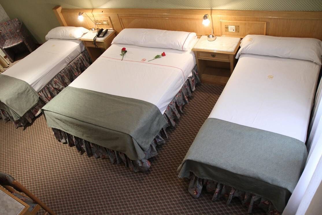 A hotel room with three single beds, each with a bedside table and lamp. The room has a checkered floor and wooden headboard.