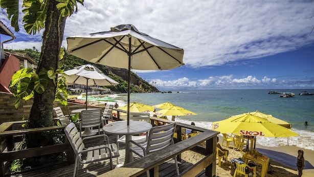 Un restaurante frente a la playa con terraza, mesas y sillas, y sombrillas amarillas.