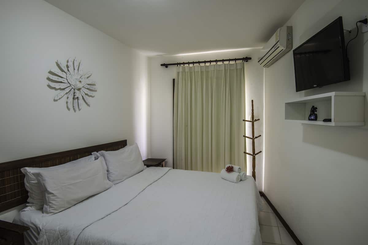 Cama bem arrumada com lençóis brancos, TV fixada na parede e janela com cortinas.