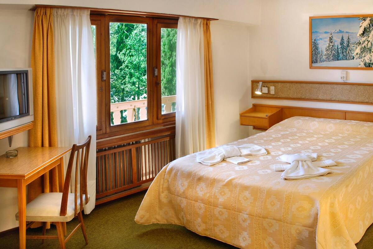 A hotel room with a bed, a desk with a chair, a TV, and a painting on the wall.