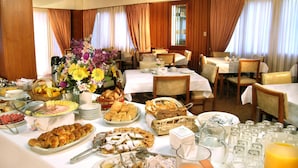 Un ricco buffet per la colazione con diverse pietanze, tra cui pasticceria, frutta e bevande, in una sala da pranzo luminosa con arredi in legno e tende.